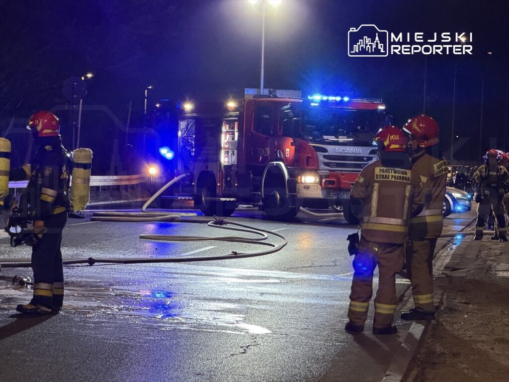 Strażacy w hełmach rozmawiają obok wozu strażackiego, na drodze leżą węże gaśnicze, w tle migają niebieskie światła.