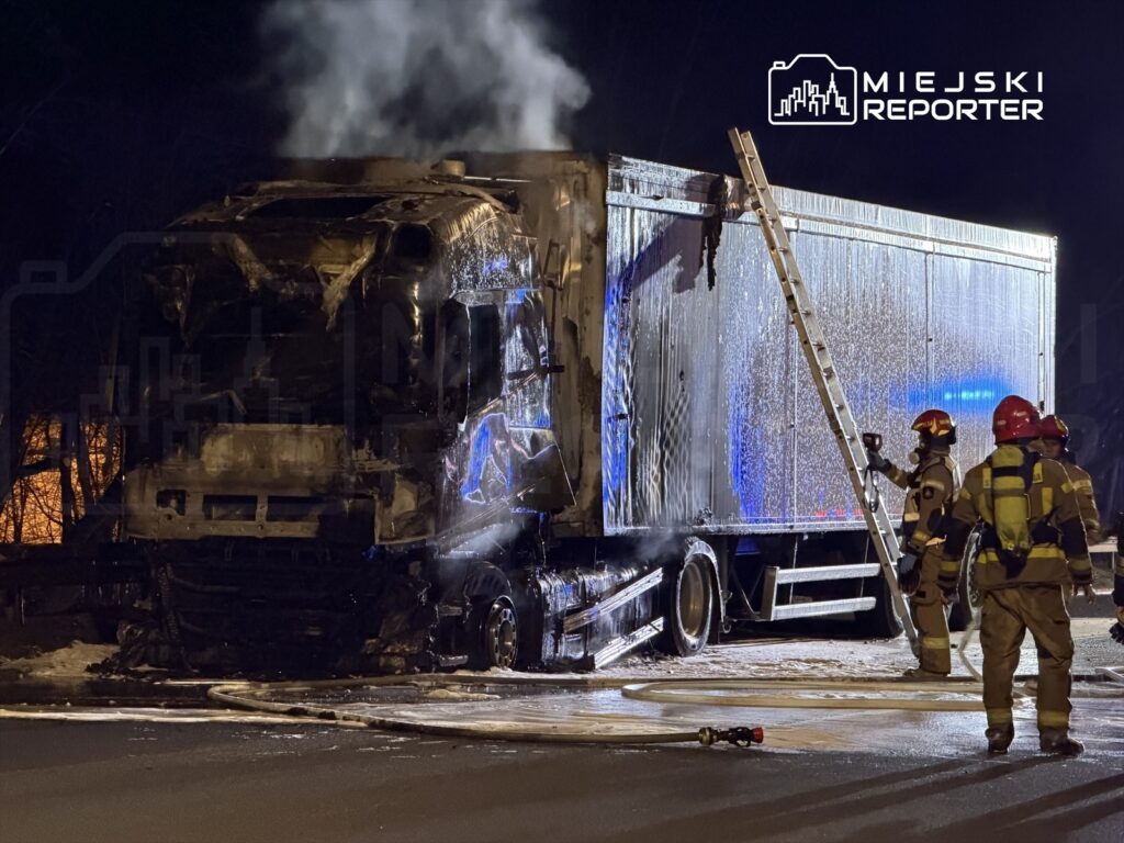 Strażacy gaszą płonącą ciężarówkę, która jest częściowo zniszczona, w nocy przy oświetlonych drzewach.