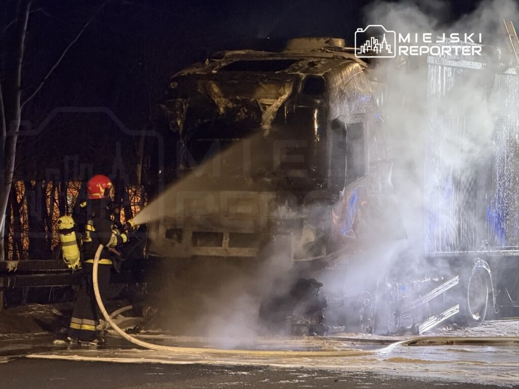 Strażak w hełmie gasi pożar ciężarówki, która stoi w dymie na drodze w nocy.