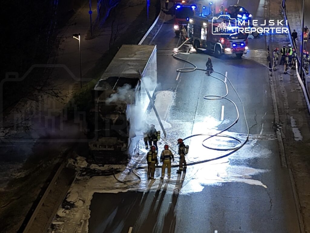 Strażacy gaszą pożar ciężarówki na drodze, obok wozów strażackich i sprzętu gaśniczego.