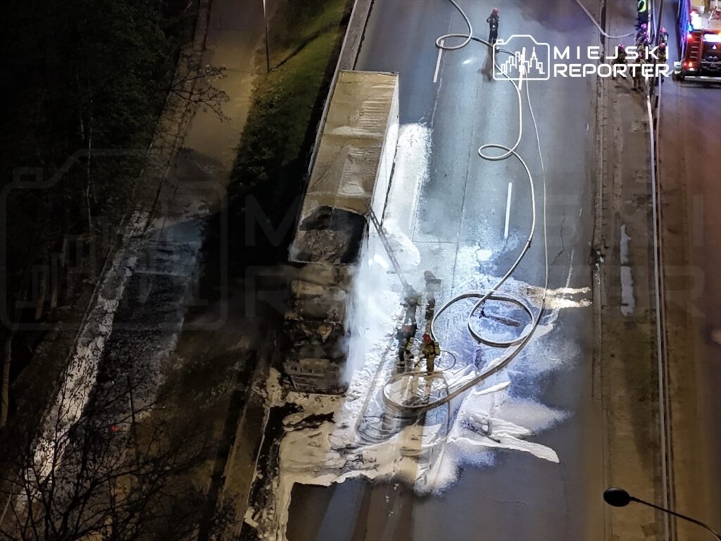 Strażacy gaszą pożar ciężarówki na jezdni, obok leży wąż gaśniczy oraz biała piana. W tle widać latarnie uliczne.