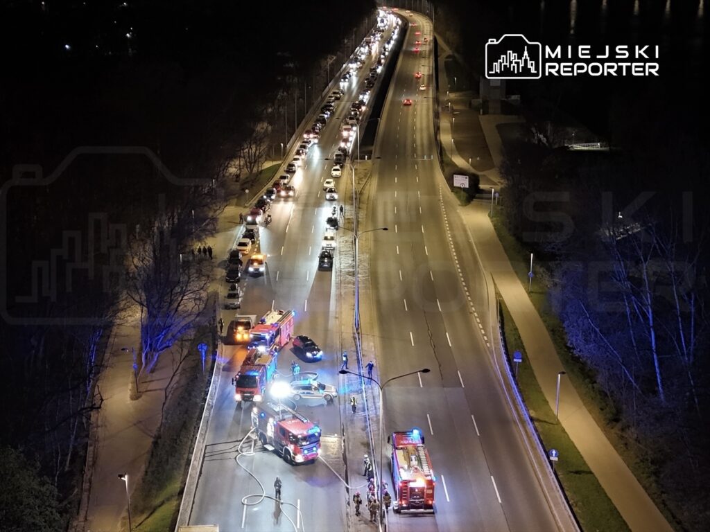 Na drodze w nocy widać pojazdy straży pożarnej oraz policyjny radiowóz, obok grupę ludzi i zablokowany ruch.