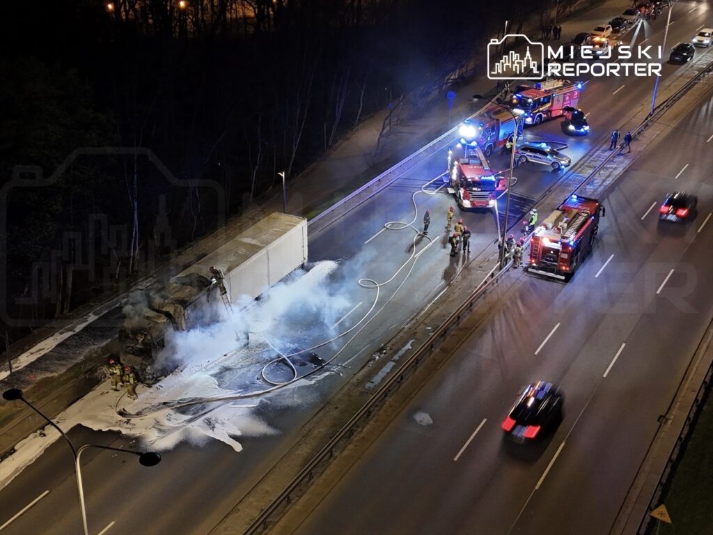 Strażacy gaszą pożar ciężarówki na jezdni, obok widać rozciągnięte węże i pojazdy ratunkowe.