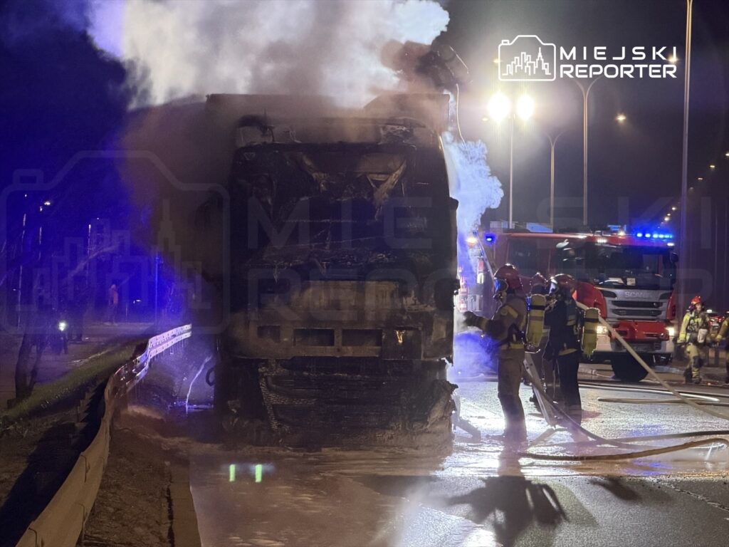 Strażacy gaszą pożar ciężarówki na drodze, obok widoczny wóz strażacki i dym unoszący się w nocy.
