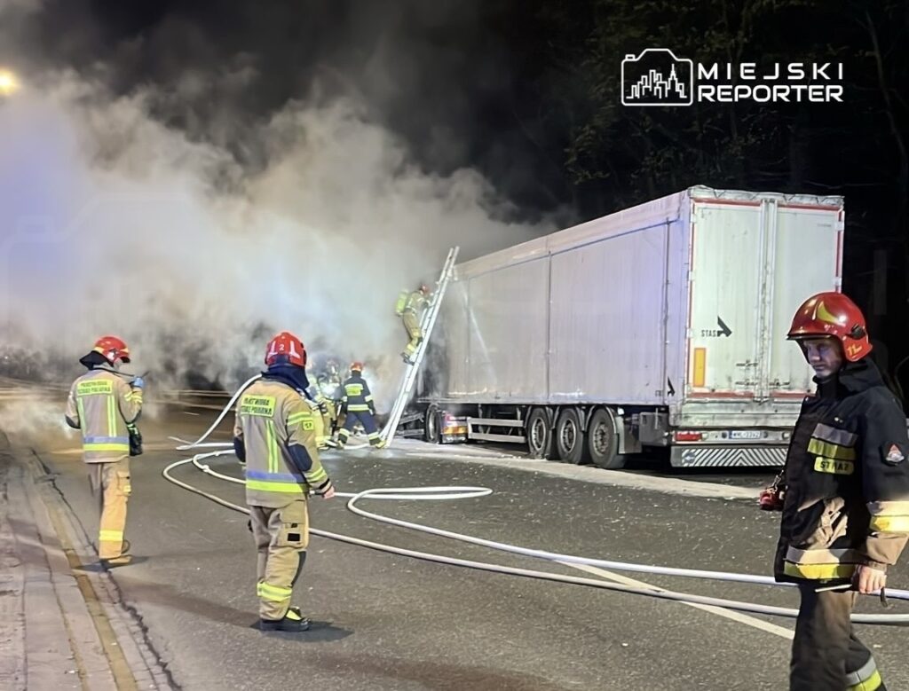 Strażacy w hełmach gaszą pożar ciężarówki, otoczeni gęstym dymem, na ulicy w nocy.