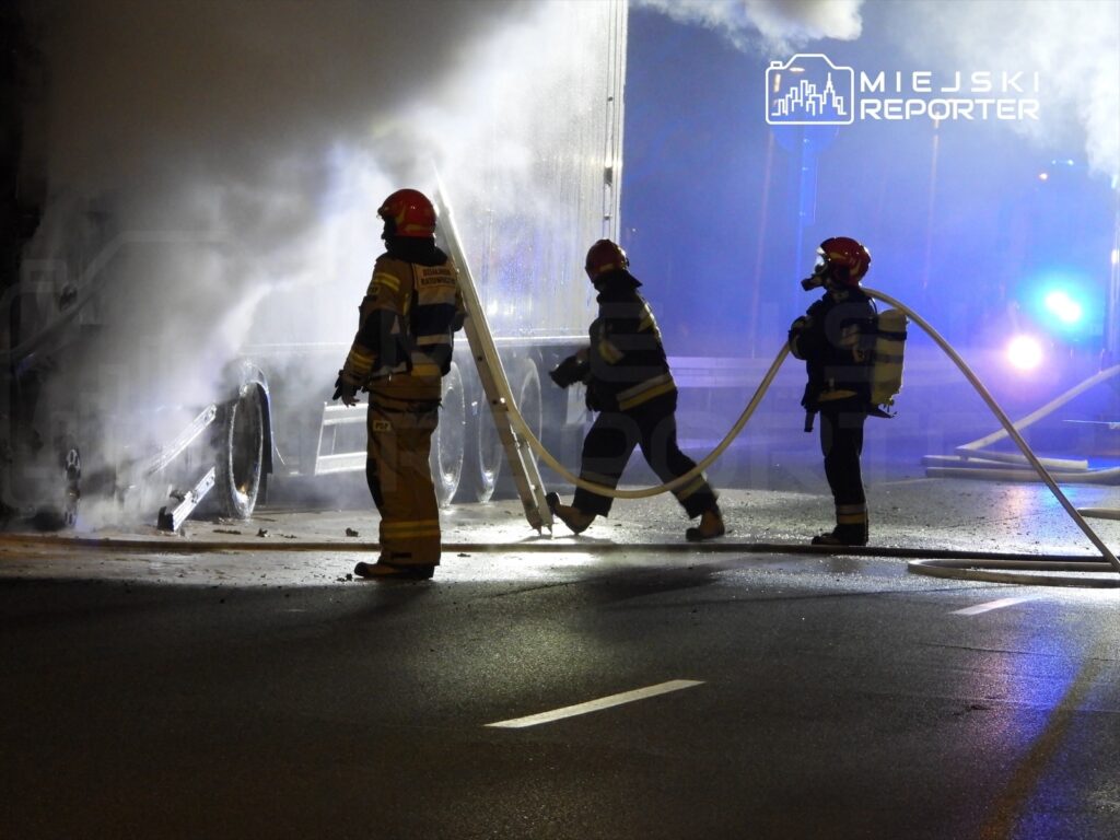 Strażacy gaszą pożar ciężarówki na drodze, otoczeni kłębami dymu i niebieskimi światłami.