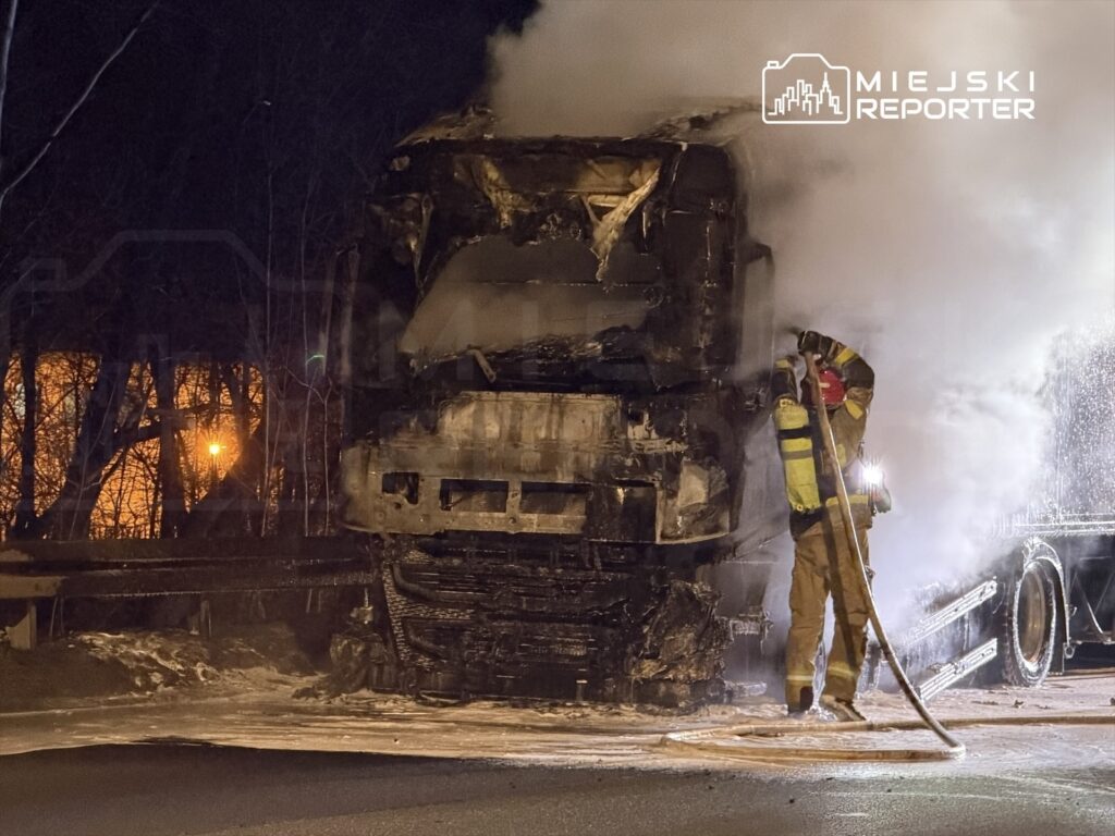 Strażak w żółtym mundurze gasi płonący samochód ciężarowy w nocy, otoczony przez dym i ciemność.
