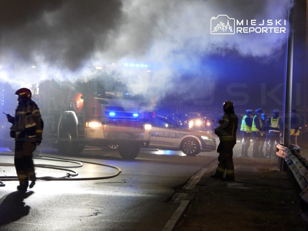 Strażacy w mundurach i kaskach oceniają sytuację w otoczeniu dymu i policyjnych świateł na ulicy.