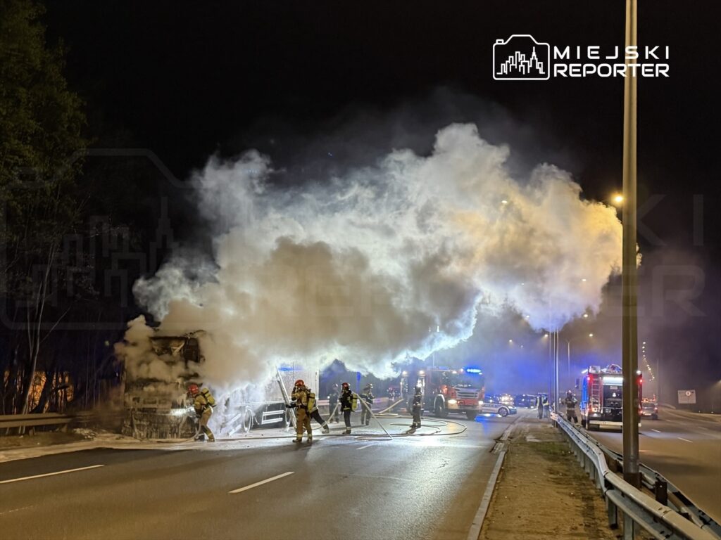 Strażacy gaszą pożar ciężarówki na drodze, otoczeni gęstym dymem w nocy, z włączonymi sygnałami świetlnymi.