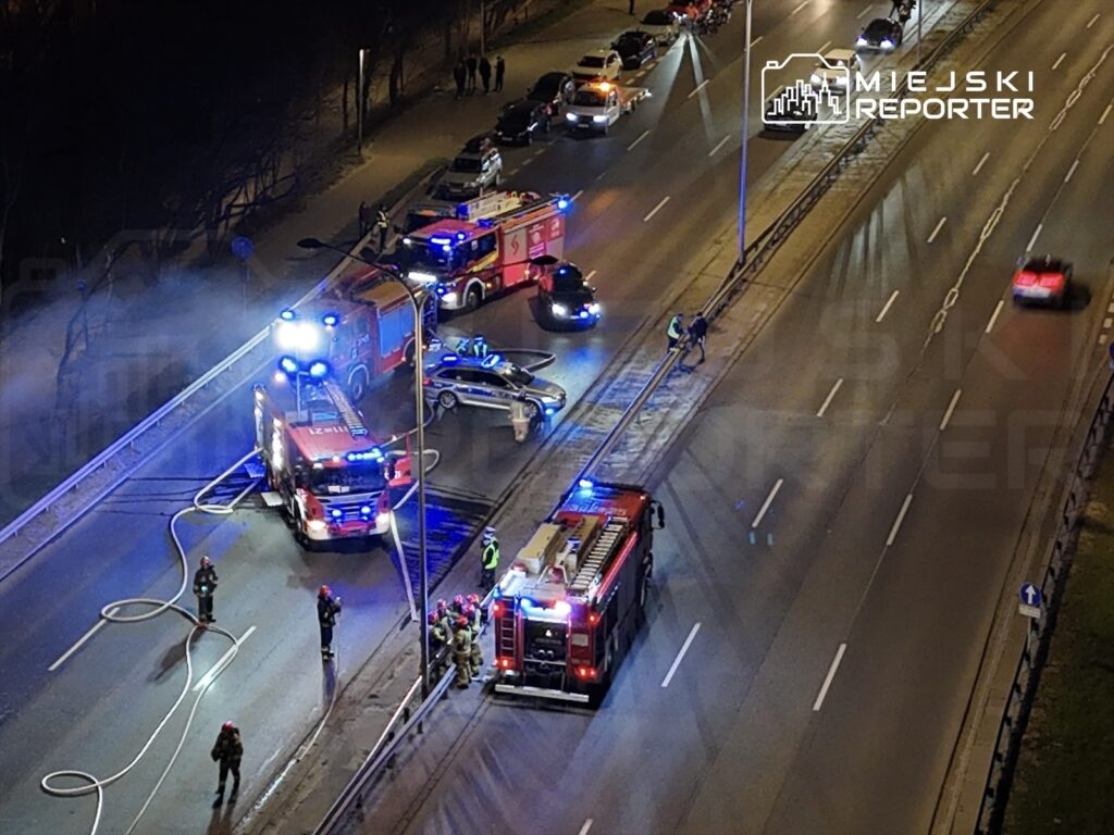 Strażacy pracują nad usunięciem skutków wypadku drogowego na ruchliwej ulicy, obok zaparkowanych pojazdów.