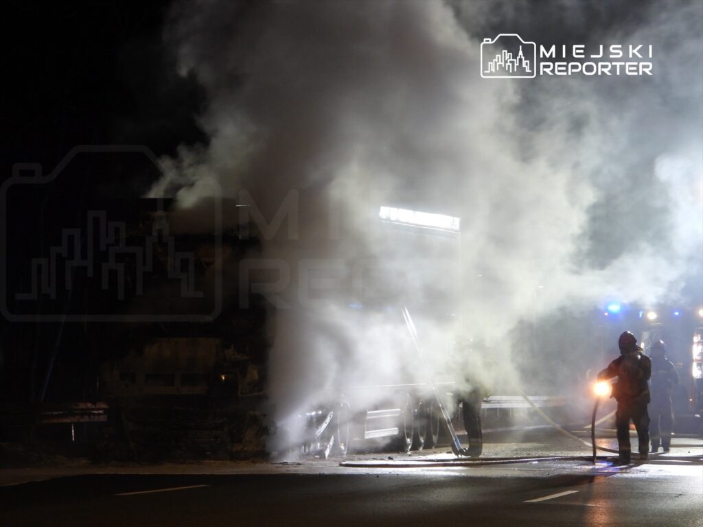 Strażacy gaszą pożar ciężarówki, otoczeni gęstym dymem na nocnej drodze.