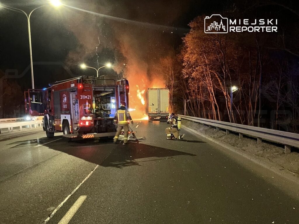 Strażacy gaszą ogień wydobywający się z ciężarówki na drodze, w otoczeniu drzew i nocnego nieba.