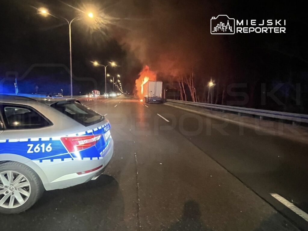 Policyjny radiowóz z włączonymi sygnałami stoi na drodze, obok płonącego ciężarowego pojazdu.