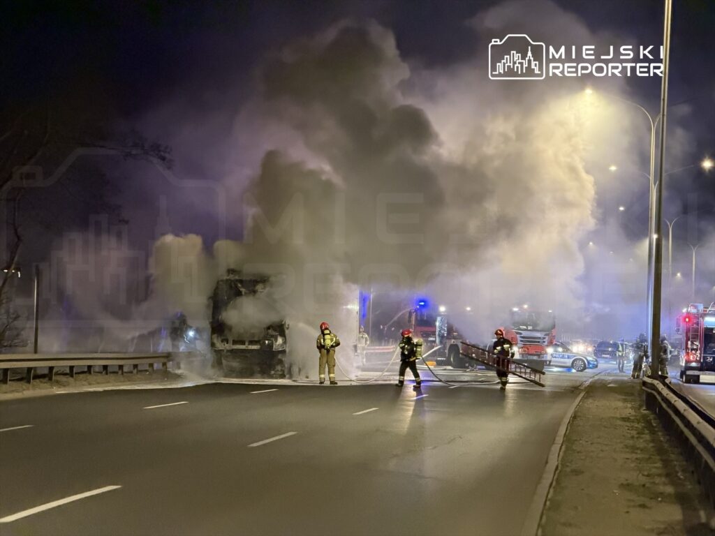Strażacy gaszą pożar ciężarówki na drodze, w otoczeniu gęstego dymu i w nocy. W tle widoczne pojazdy ratunkowe.