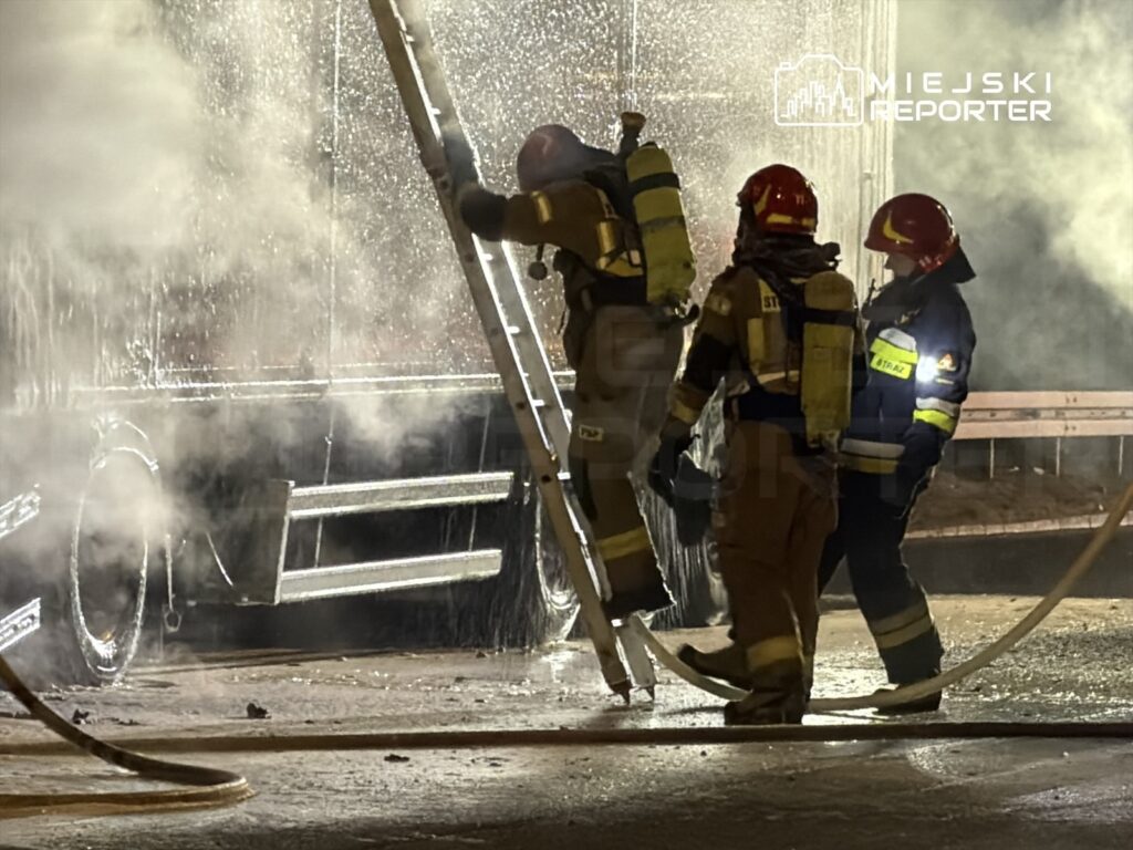 Strażacy w hełmach i mundurach gaszą pożar przy użyciu węża strażackiego, otoczeni przez dym i wodę.