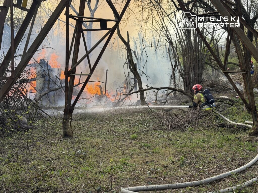 Strażak w kasku i mundurze gasi ogień, który strzeże zarośnięte tereny w pobliżu zniszczonej konstrukcji.