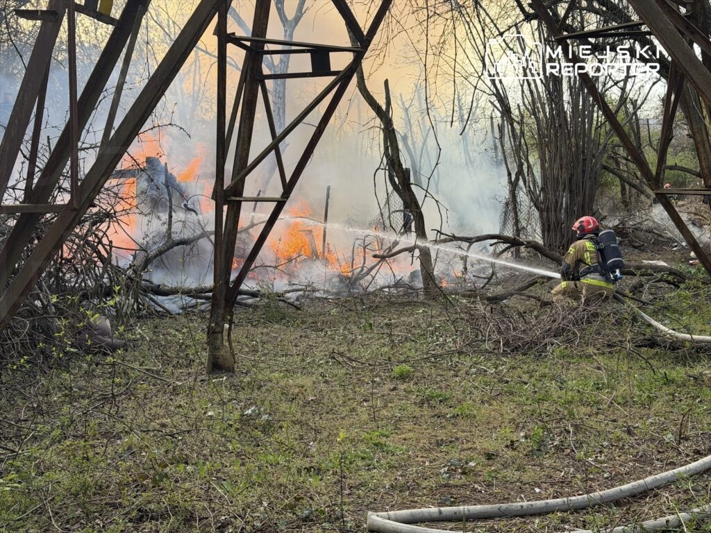 Strażak w hełmie gasi ogień w zaroślach, otoczony dymem i płomieniami, w tle widoczne metalowe konstrukcje.