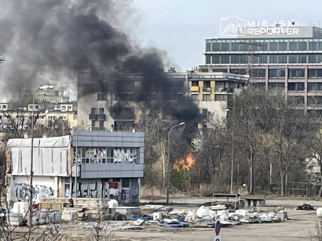 Czarny dym unoszący się z płonącego budynku w miejskim otoczeniu, w tle nowoczesne biurowce.
