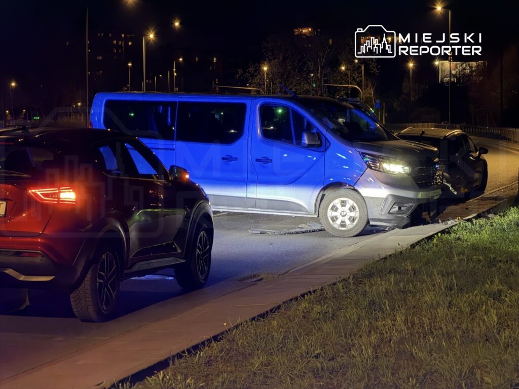 Na drodze nocą zaparkowane są dwa samochody osobowe i jeden van, w tle widać oświetlone ulice i budynki.