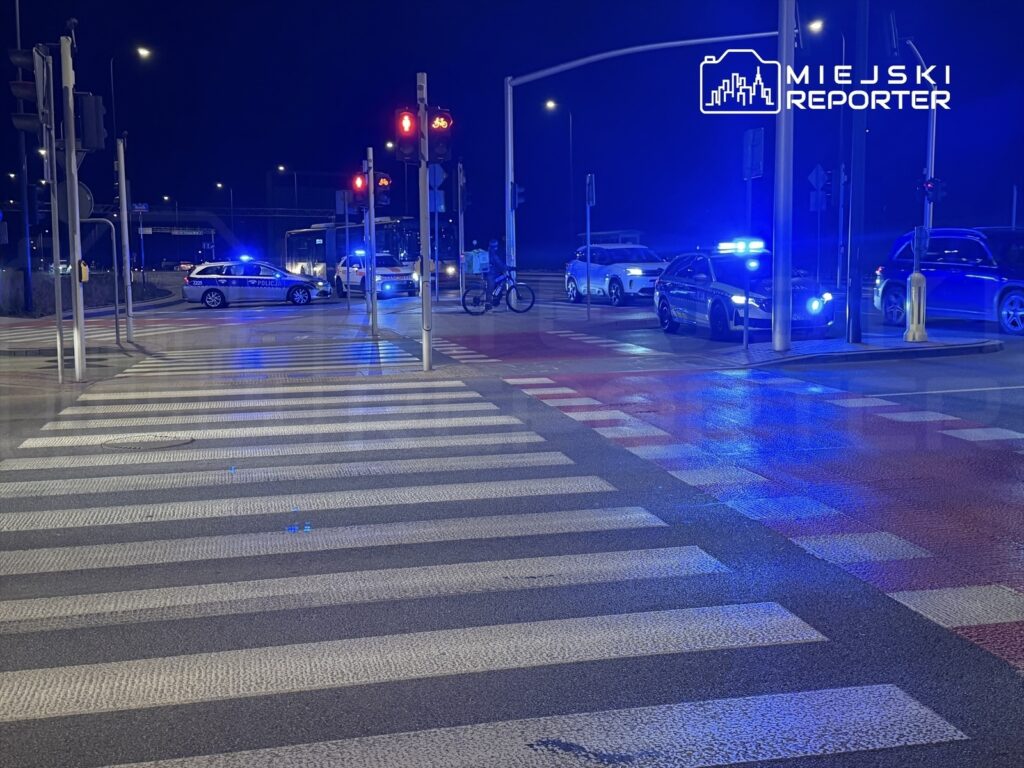 Policyjny radiowóz z włączonymi sygnałami świetlnymi zatrzymany na przejściu dla pieszych w nocy.