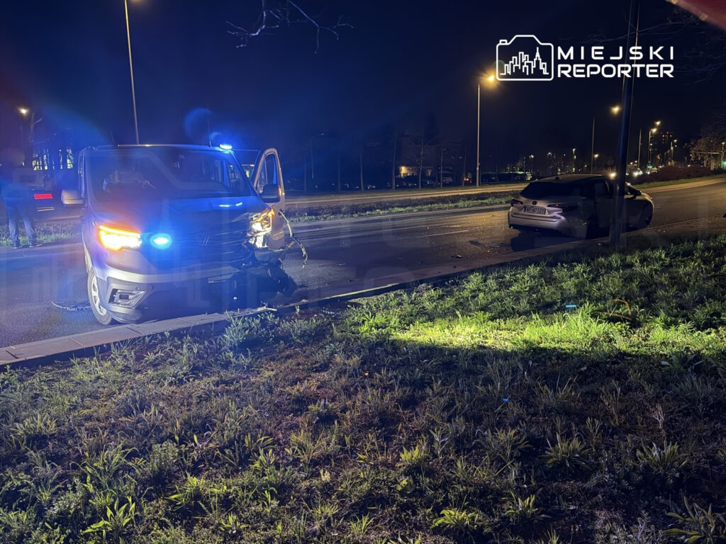 Policyjny samochód z włączonymi sygnalizatorami świetlnymi stoi obok uszkodzonego pojazdu na oświetlonej ulicy.