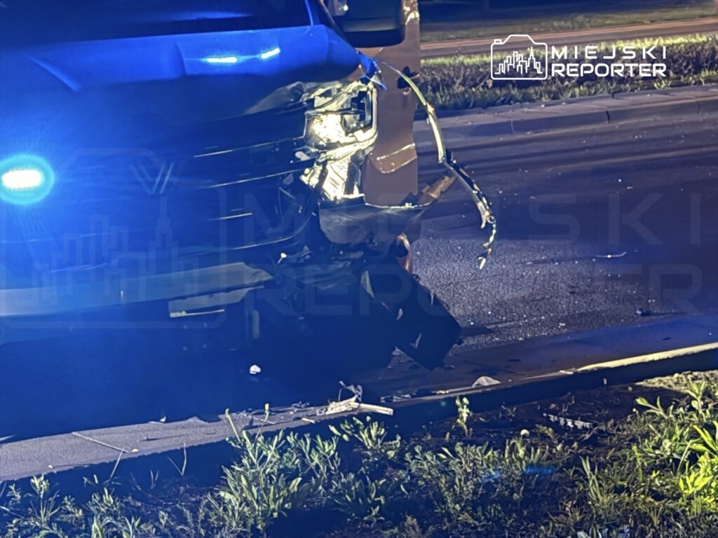 Uszkodzony pojazd z włączonymi światłami policyjnymi stoi na jezdni, otoczony przez trawę i zniszczone fragmenty.