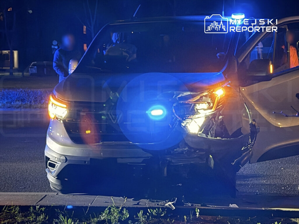 Uszkodzony pojazd policyjny z uszkodzoną przednią częścią, zatrzymany na drodze w nocy, otoczony niebieskimi światłami.