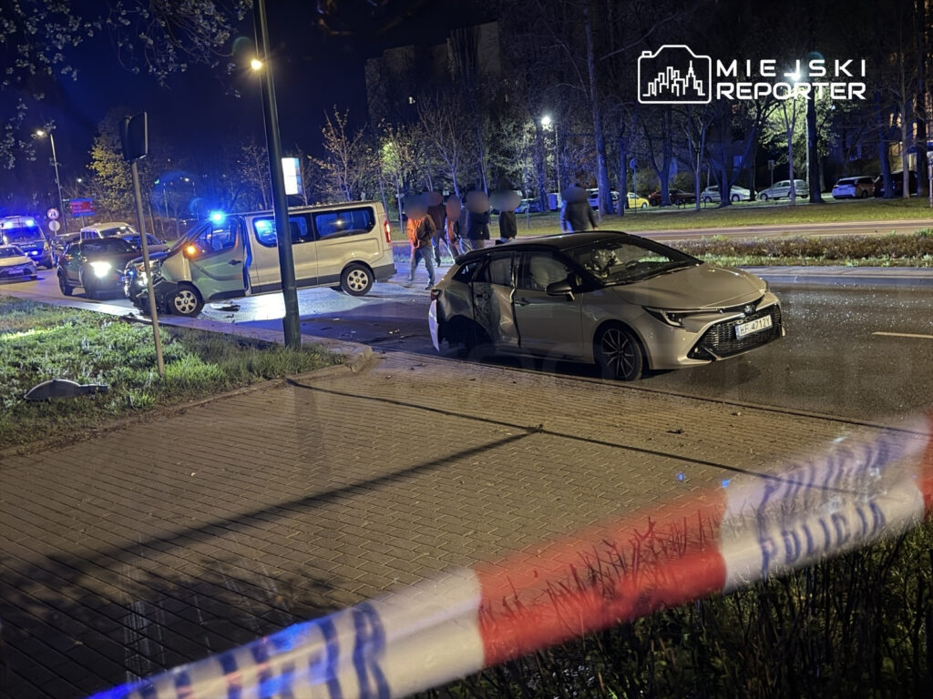 Na ulicy nocą, dwa uszkodzone samochody po kolizji, wokół policyjne radiowozy i zebrani świadkowie.