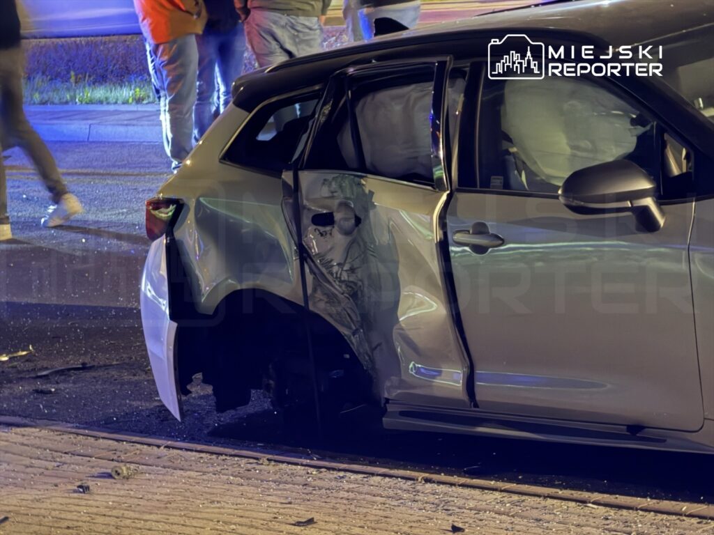 Uszkodzony samochód osobowy z wgniecioną boczną częścią, obok grupka ludzi w tle, w otoczeniu policyjnych świateł.