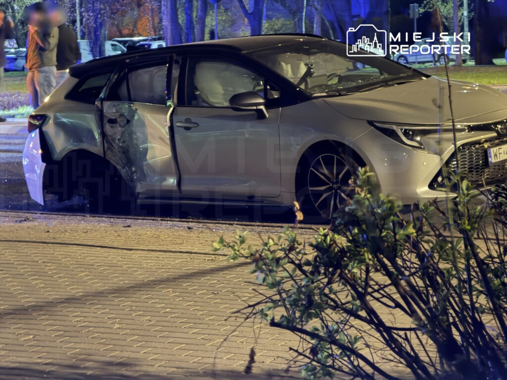 Uszkodzony samochód osobowy z wgniecioną tylną częścią stoi na chodniku, a w tle widać zebranych ludzi.