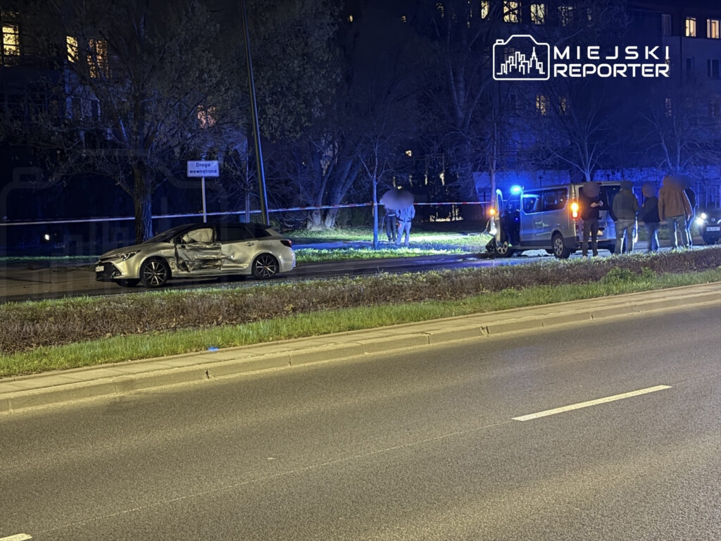 Uszkodzony samochód osobowy stoi na drodze, a obok grupka osób oraz pojazd policyjny z włączonymi sygnałami świetlnymi.
