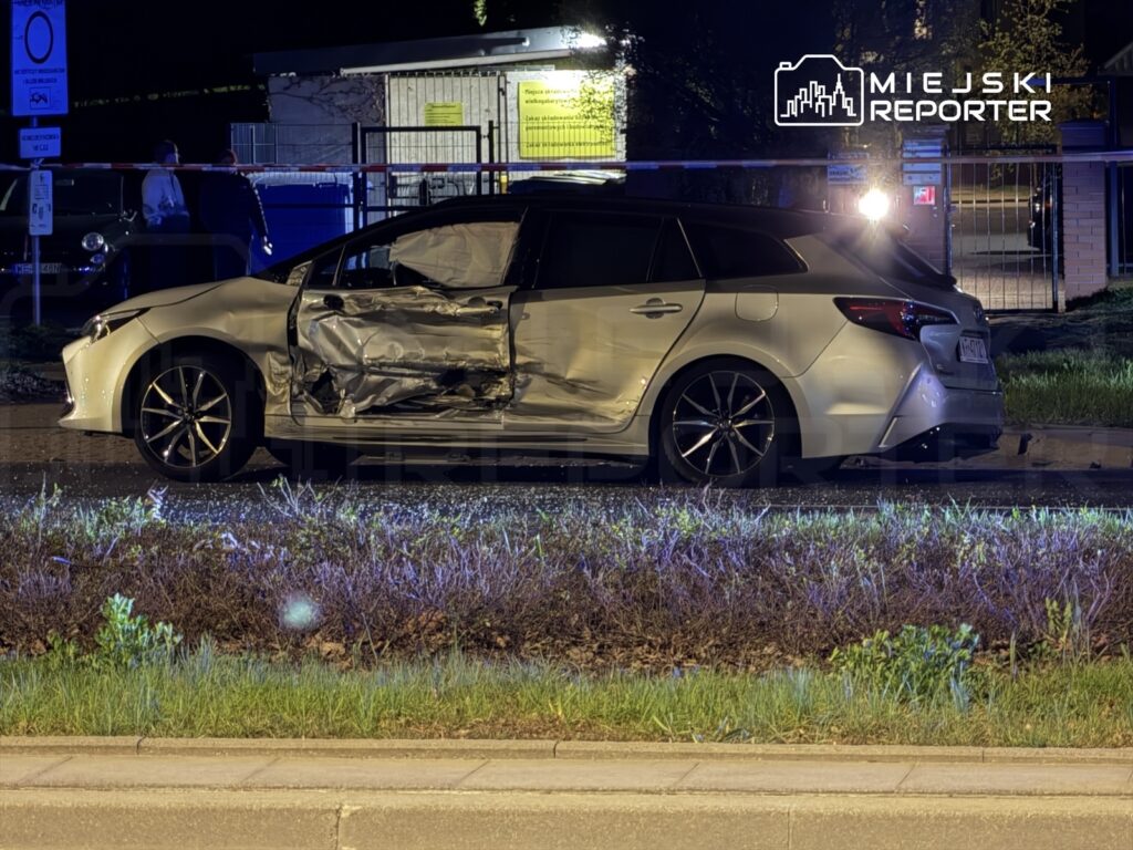 Uszkodzony samochód osobowy z wgniecioną boczną częścią stoi na poboczu drogi, w tle widoczne są policyjne światła.