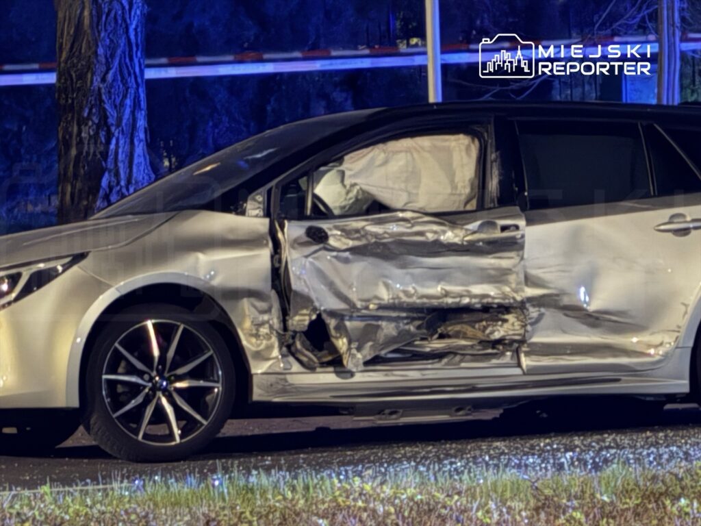 Uszkodzony samochód osobowy z wgniecioną lewą stroną stoi na poboczu drogi, otoczony niebieskim światłem policyjnym.