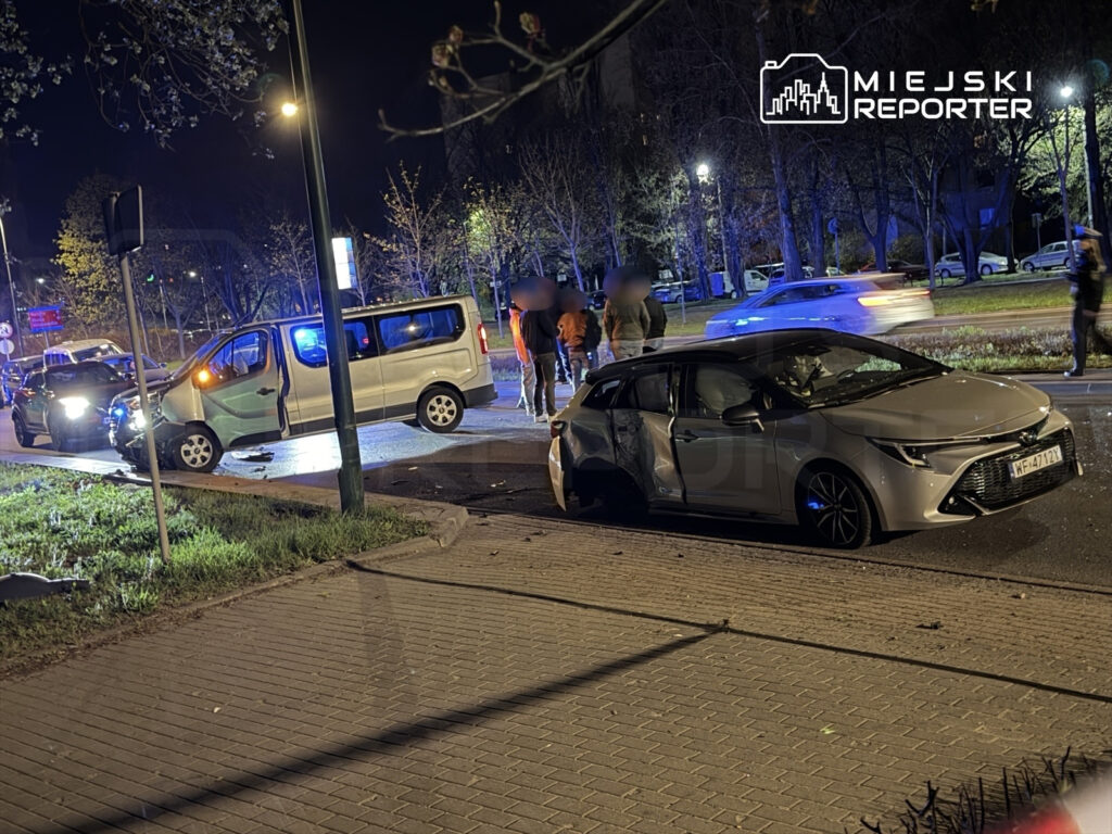 Na drodze nocą doszło do wypadku, w którym białe auto dostawcze z uszkodzoną przednią częścią stoi na poboczu, a obok grupka ludzi obserwuje sytuację.