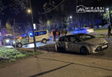 Na drodze nocą doszło do wypadku, w którym białe auto dostawcze z uszkodzoną przednią częścią stoi na poboczu, a obok grupka ludzi obserwuje sytuację.