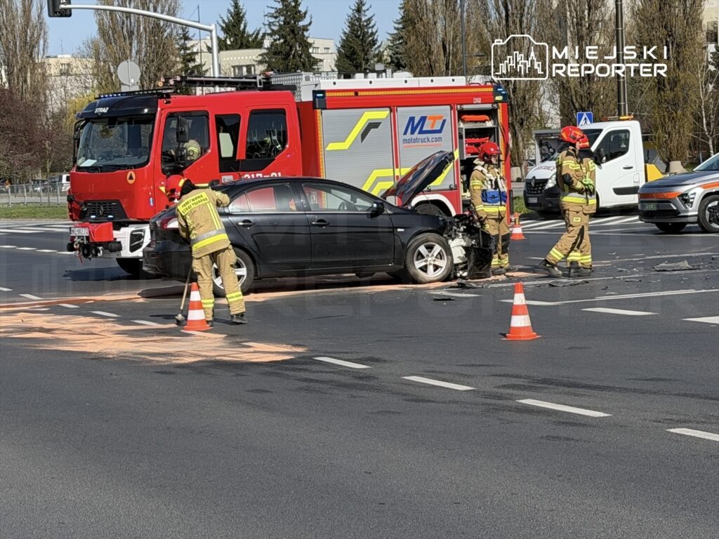 Strażacy usuwają rozbite auto na skrzyżowaniu, obok stoi wóz strażacki, a na jezdni leży plama płynów.