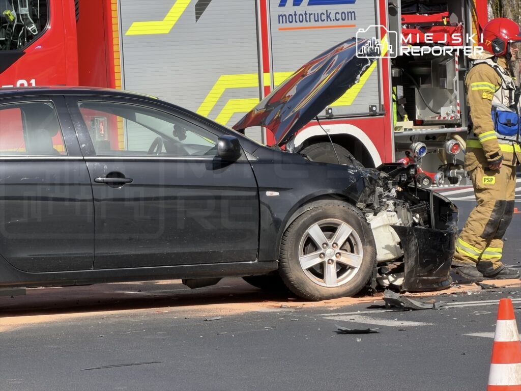 Czarny samochód osobowy z uszkodzoną przednią częścią stoi na drodze, obok strażak w mundurze z wozem strażackim w tle.
