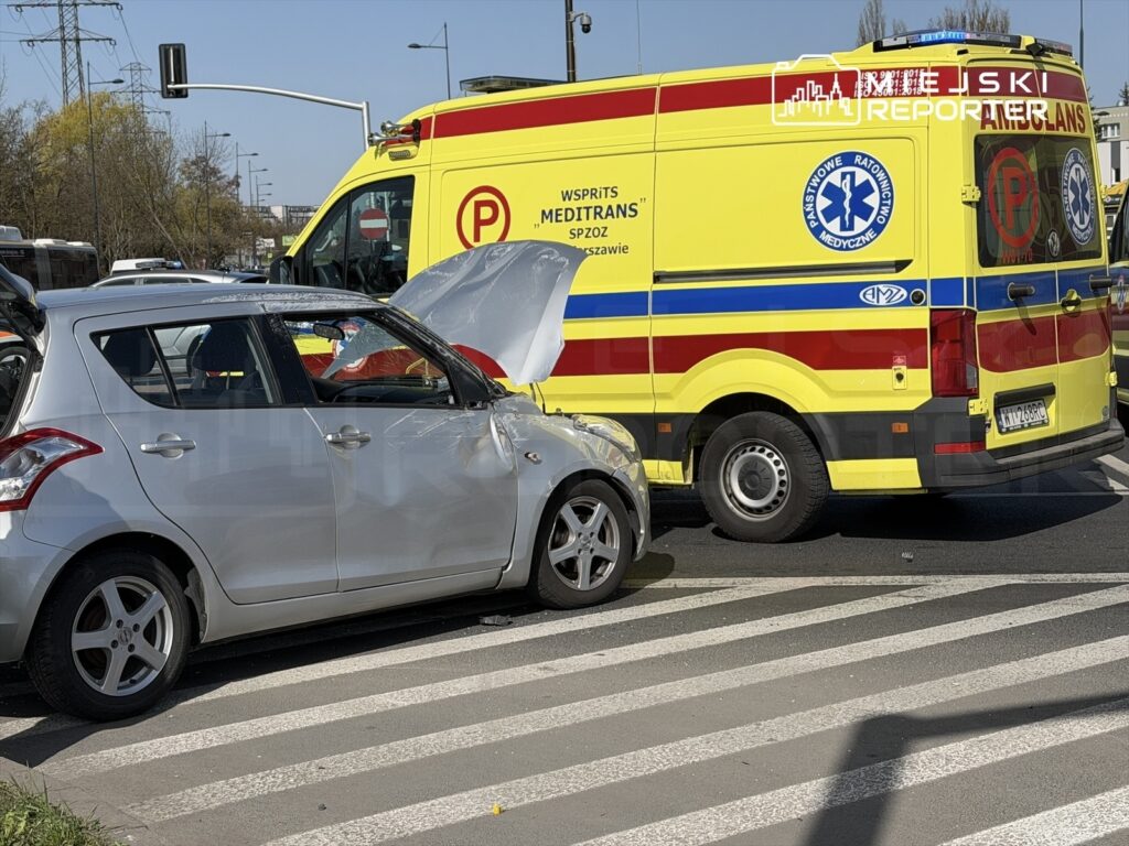 Srebrny samochód osobowy z uszkodzoną maską stoi na pasach, obok niego zatrzymana karetka pogotowia.