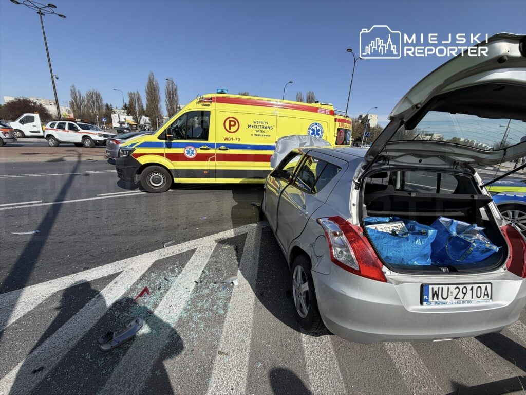 Ambulans z włączonymi sygnałami zatrzymany obok uszkodzonego samochodu osobowego na ruchliwej ulicy. W tle widać inne pojazdy.