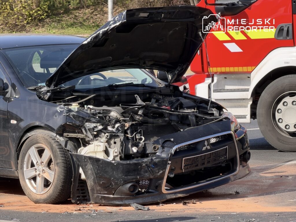 Uszkodzony czarny samochód osobowy z otwartą maską stoi na drodze, obok widać wóz strażacki.