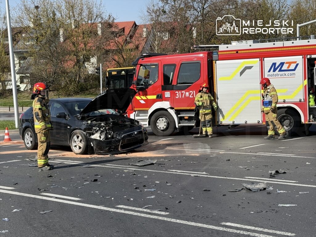 Strażacy w mundurach interweniują przy uszkodzonym samochodzie osobowym na drodze, obok stoi wóz strażacki.