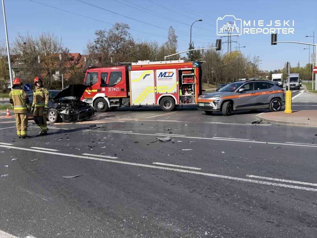 Strażacy rozmawiają obok zderzonego samochodu osobowego na skrzyżowaniu, obok stoi wóz strażacki.
