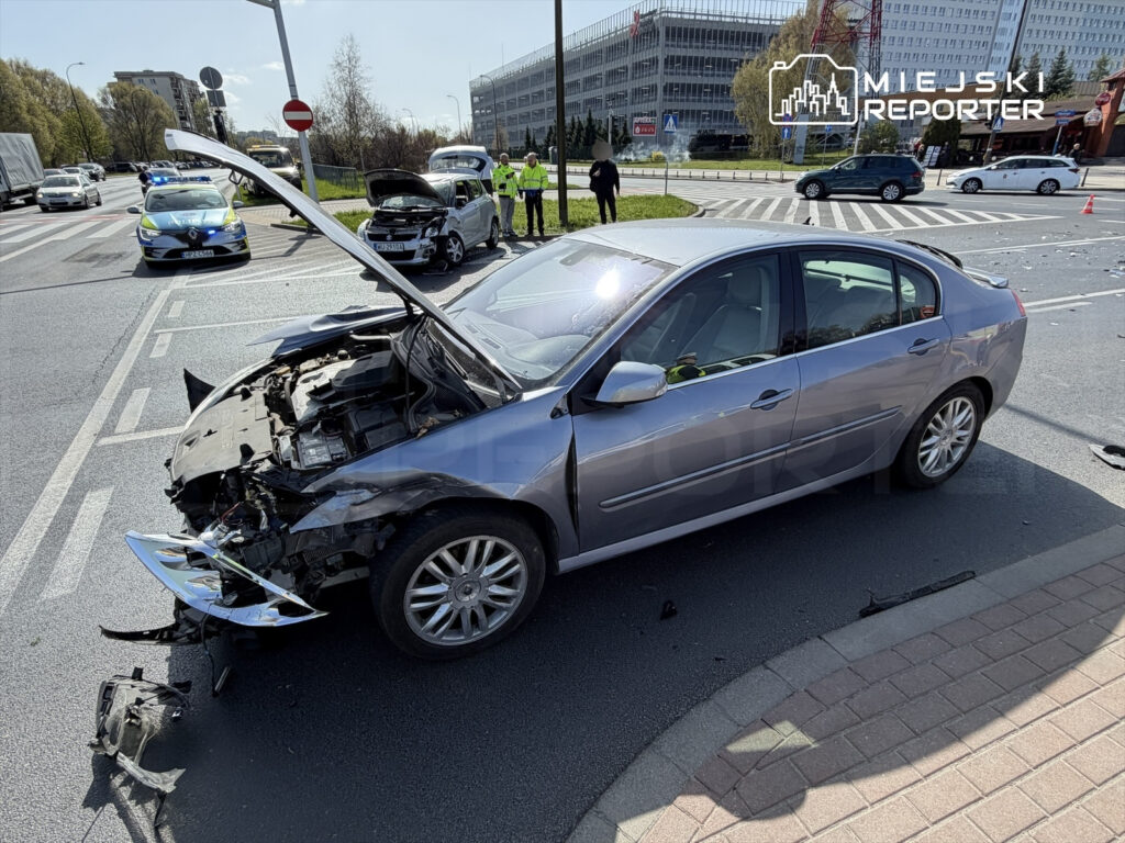 Uszkodzony samochód osobowy z otwartą maską stoi na skrzyżowaniu, obok policjanci zabezpieczają miejsce wypadku.