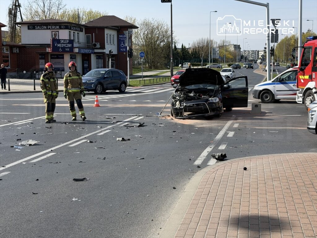 Dwaj strażacy w mundurach stoją obok uszkodzonego samochodu na drodze, wokół leżą fragmenty wraku i ślady wypadku.