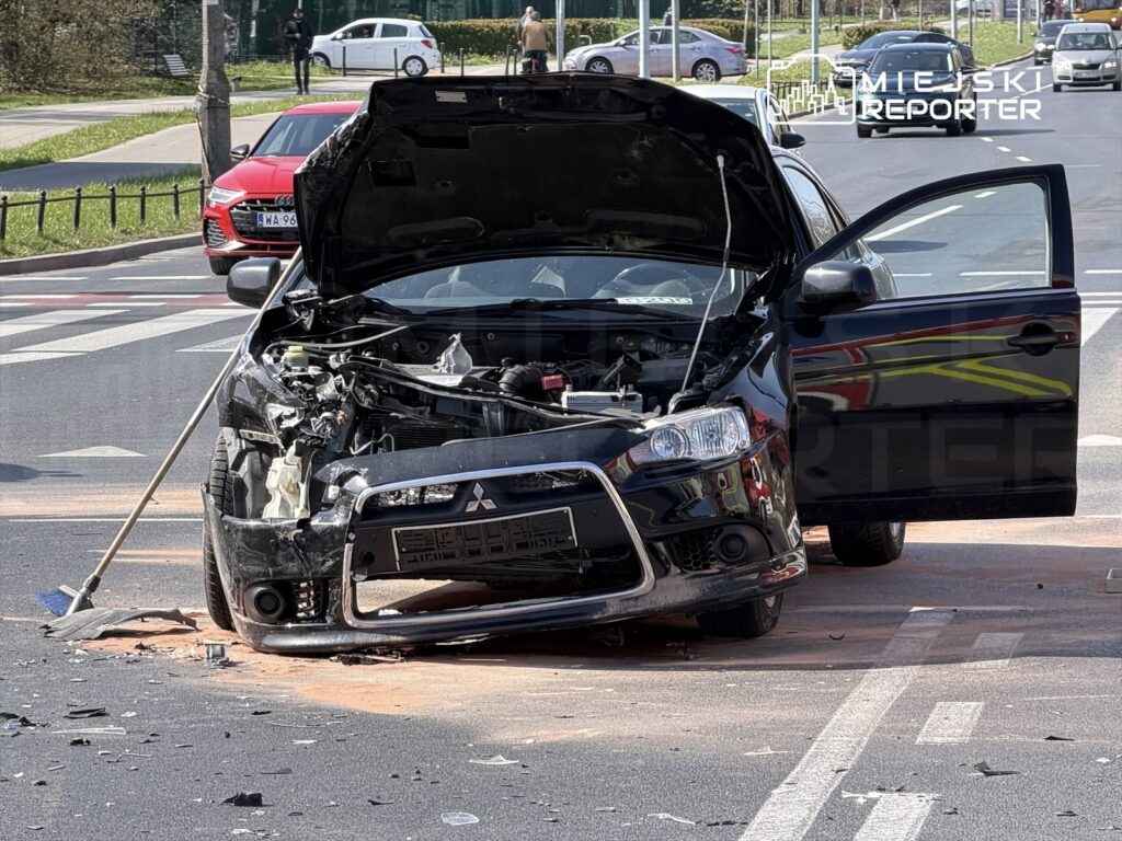Zniszczony czarny samochód osobowy z otwartą maską stoi na drodze, obok leży miotła i pozostałości wypadku.