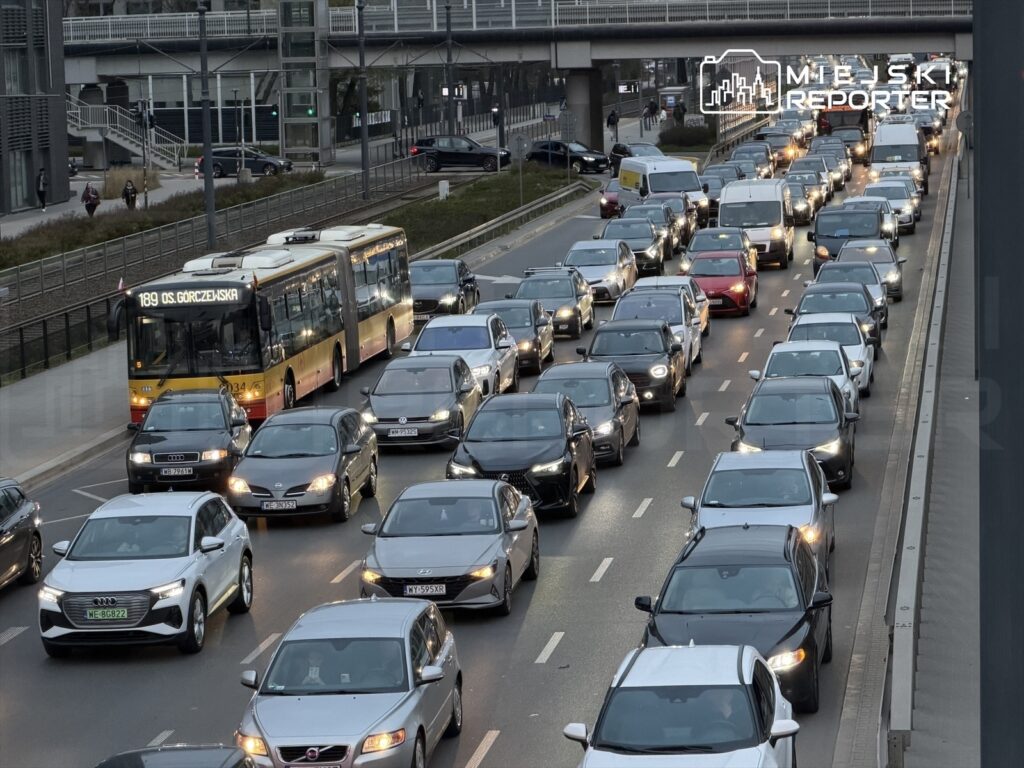 Zatłoczona ulica z wieloma samochodami osobowymi i autobusem komunikacji miejskiej, w tle widoczny most.