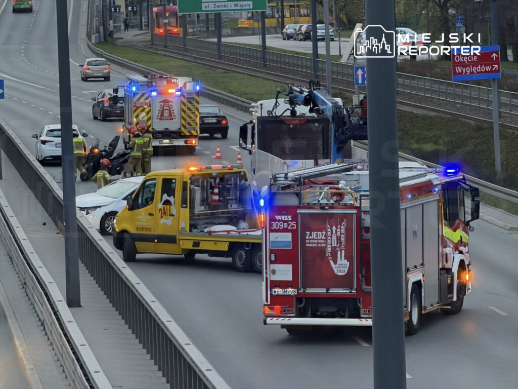 Na jezdni doszło do wypadku, w którym uczestniczyły dwa samochody oraz motocykl. Strażacy i ratownicy medyczni udzielają pomocy poszkodowanym. W tle widoczne inne pojazdy oraz znaki drogowe.