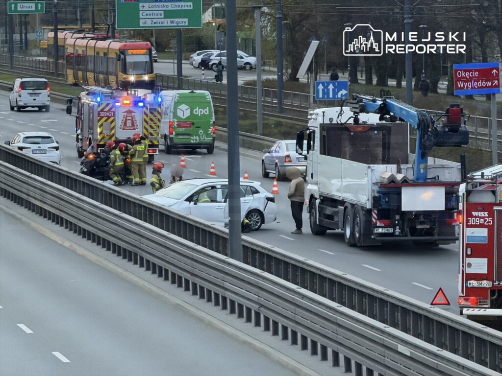 Grupa strażaków udziela pomocy przy wypadku drogowym na ruchliwej jezdni, obok stoi pojazd strażacki.