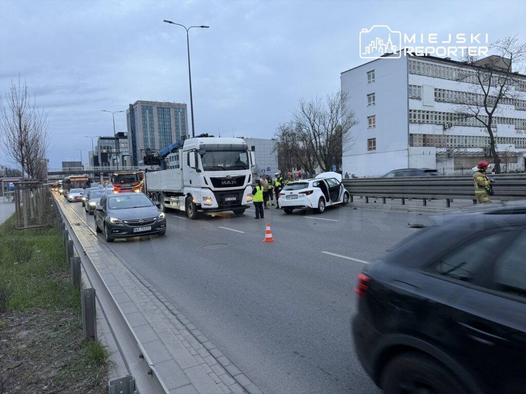 Wielu ratowników i policjantów zabezpiecza miejsce wypadku drogowego na ruchliwej ulicy z uszkodzonym samochodem osobowym.