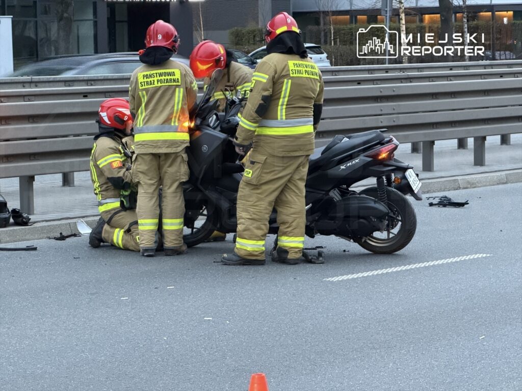 Czterech strażaków w kaskach i mundurach pomaga poszkodowanemu przy przewróconym skuterze na jezdni.